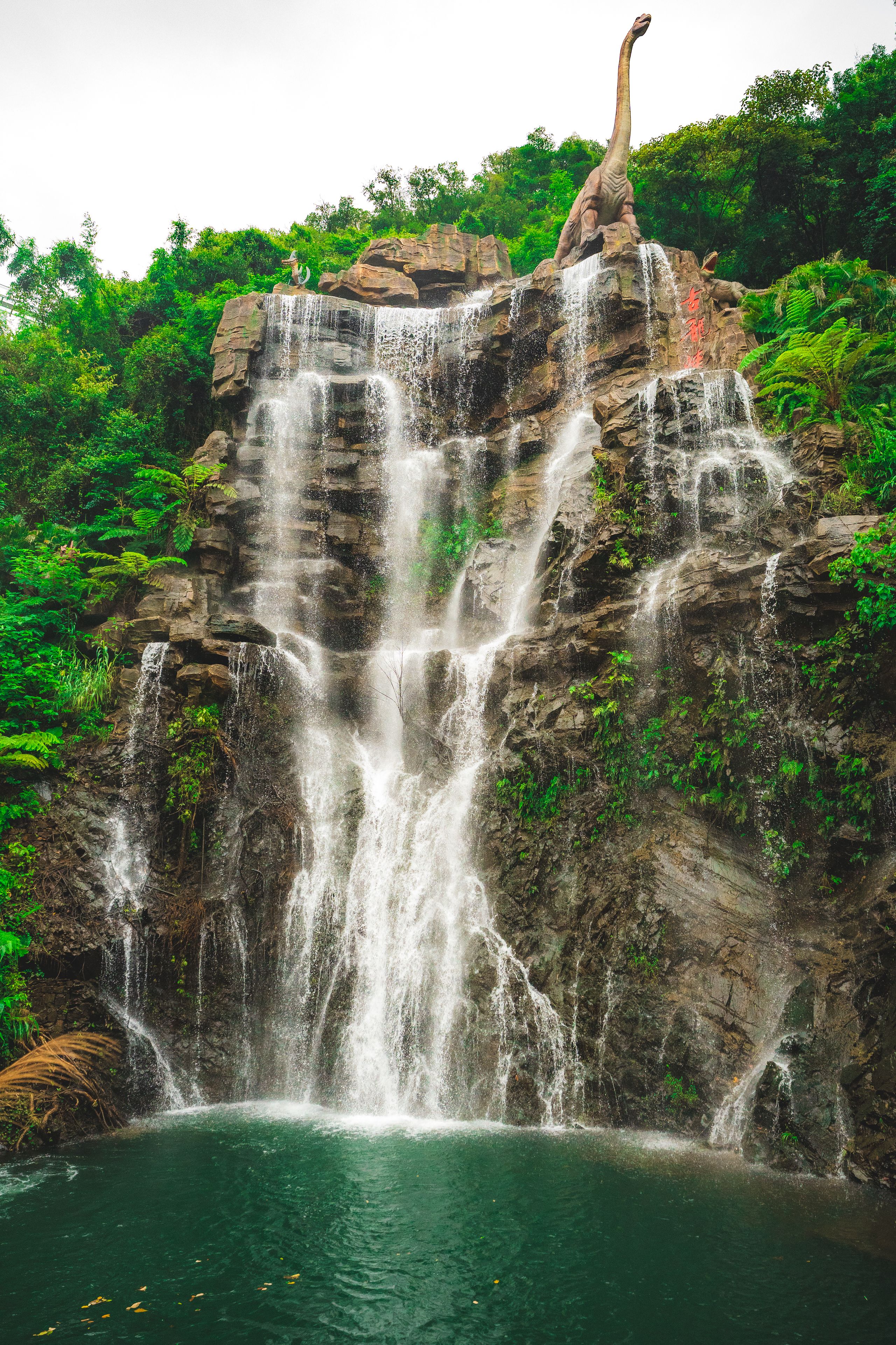 Wasserfall über Felsen unter einer Dinosaurierstatue in einem grünen Park